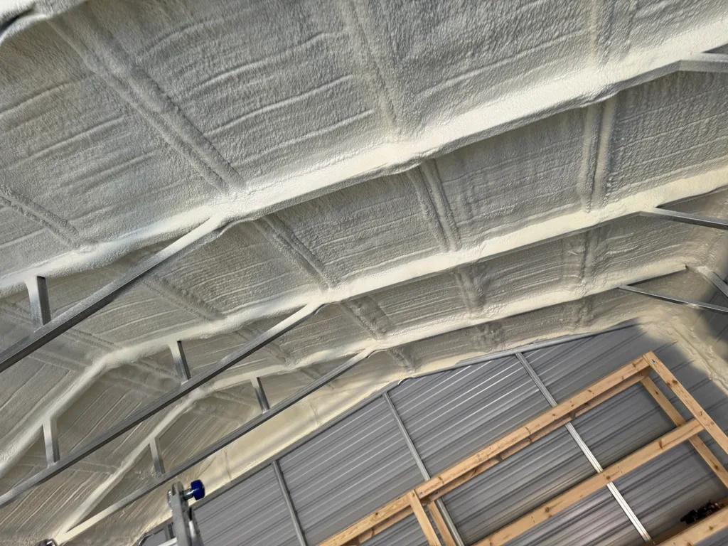 Pale foam sprayed across a steel building roof with purlins running the full span