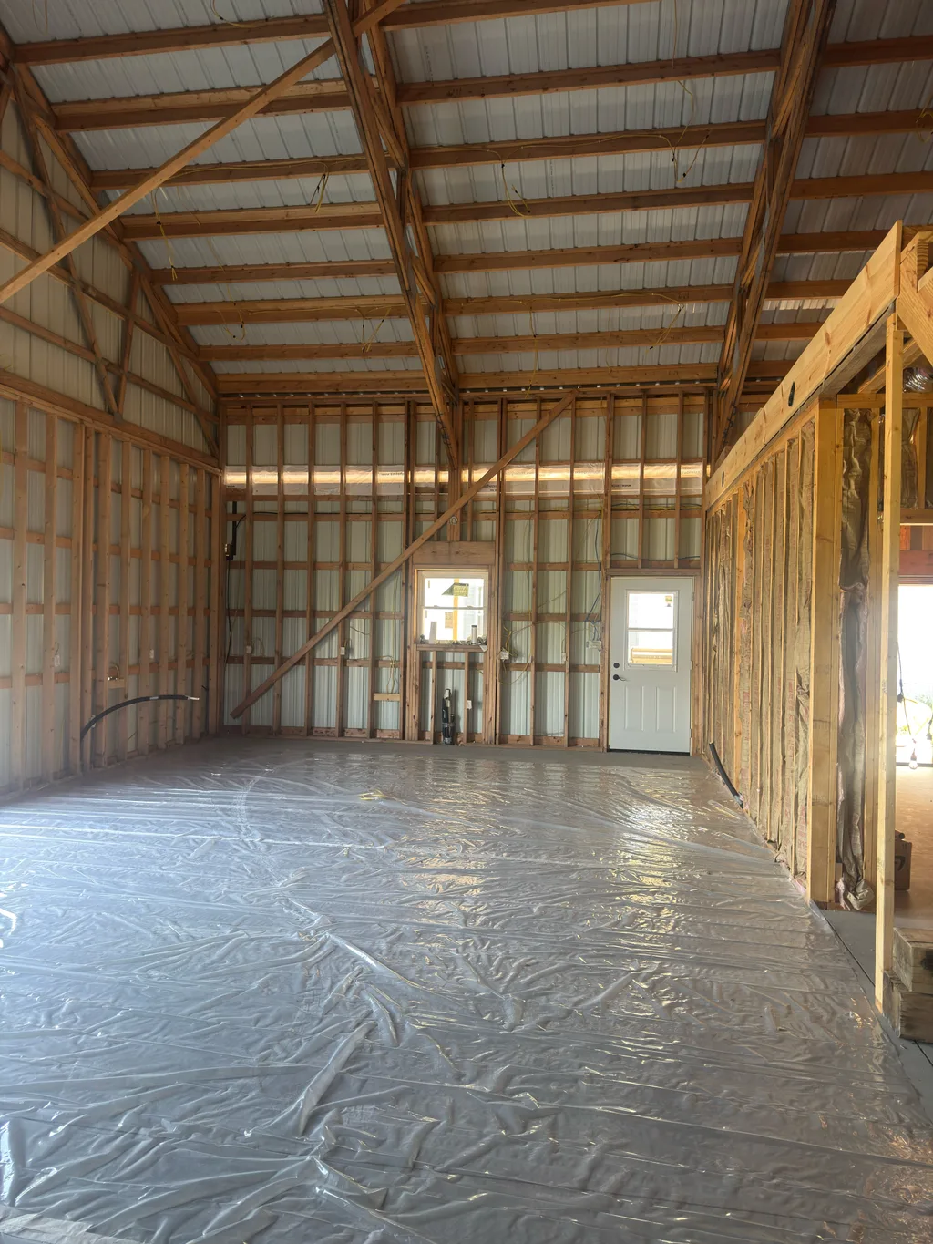 Pole barn framing with plastic sheeting over the floor and a white door at the far end
