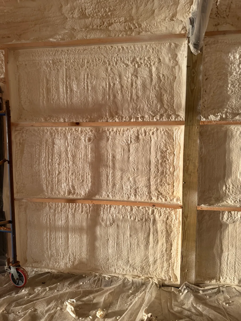 Cream foam sprayed across a wall between timber girts with pipework at the corner